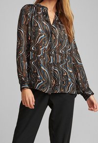 Blusa a fantasia fluida in nero, marrone e blu, con chiusura a bottoni frontale, leggermente a sbuffo e realizzata in un tessuto morbido e leggero.