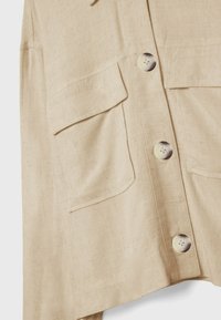 Gros plan sur une chemise en lin beige avec de grandes poches à rabat et quatre boutons ronds de couleur claire sur le devant.