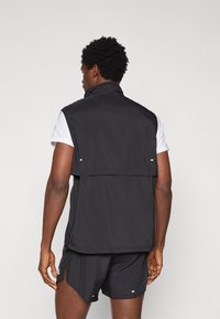 Μαύρο γιλέκο χωρίς μανίκια από ελαφρύ ύφασμα με ψηλό γιακά, διαθέτει πτερύγιο στην πλάτη και τσέπες με φερμουάρ στα πλαϊνά, φορεμένο πάνω από λευκό πουκάμισο.