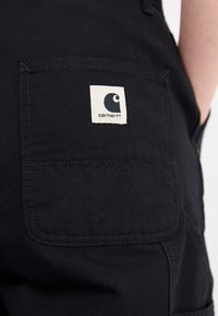 Des salopettes en tissu noir présentent une grande poche arrière avec une étiquette carrée blanche affichant le logo de la marque. Le tissu a une texture lisse.