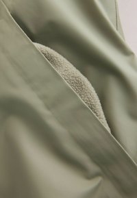 Materialul exterior verde, cu o textură netedă, înconjoară o căptușeală interioară crem, pufoasă, evidențiind o combinație de materiale contrastante.