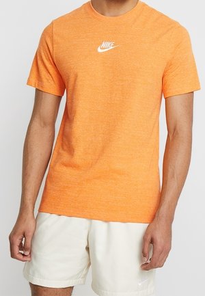 Un homme porte un t-shirt Nike orange vif et un short Nike beige clair, debout avec les bras détendus le long du corps.