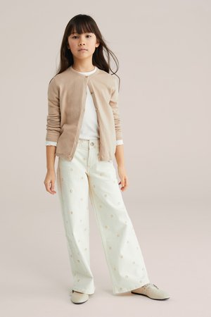 Jong meisje dat een beige vest draagt, een wit shirt, wijde witte broek met bloemmotieven en beige schoenen, staand tegen een effen achtergrond.