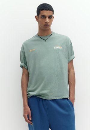 Ung mand, der står mod en hvid baggrund iført lysegrøn t-shirt med opkørte ærmer og blå bukser, med sort perlehalskæde.