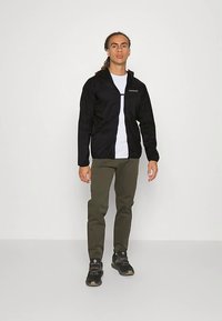 Vyras stovi vilkėjęs juodą striukę, baltą marškinėlį, alyvuogių spalvos kelnes ir juodus žygių batus prieš šviesų neutralų foną.