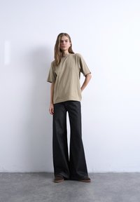 Γυναίκα που στέκεται μπροστά σε έναν λευκό τοίχο φορώντας μια μπεζ υπερμεγέθη μπλούζα, φαρδιά μαύρα τζιν και καφέ παπούτσια σε γκρι πάτωμα.