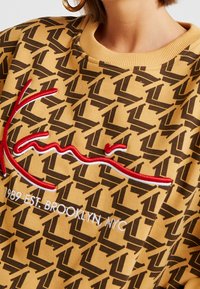 Sudadera color mostaza con un patrón geométrico oscuro, gran firma bordada en rojo y texto en blanco "1989 EST. BROOKLYN, NYC" en el frente.