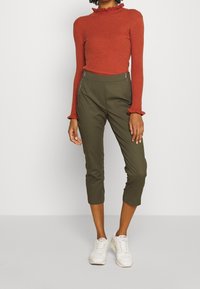 Rdzawa, prążkowana bluzka z długim rękawem i falbaną przy kołnierzyku, połączona z oliwkowymi, krótkimi spodniami z bocznymi zamkami i białymi trampkami.