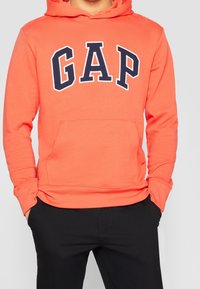 Persona que lleva una sudadera con capucha de color naranja brillante con grandes letras en azul marino "GAP" en el frente, combinada con pantalones negros, con las manos en los bolsillos.