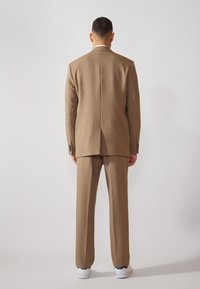 Traje marrón a medida con un blazer estructurado y pantalones a juego, combinado con zapatillas blancas. Tejido suave con una silueta limpia.