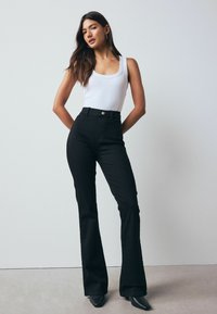 High-waisted black jeans made from denim with a slim fit and flared leg, paired with a white tank top. Simple button closure detail.