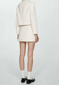 Blazer texturé blanc et mini-jupe assortie, avec un design structuré, manches longues et détail découpé dans le dos. Chaussures et chaussettes noires.