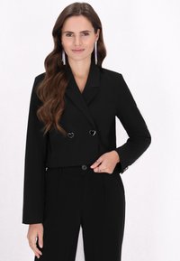 Sacou negru scurt cu butoane argintii în formă de inimă, mâneci lungi și croială ajustată, asociat cu pantaloni negri asortați.