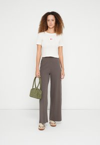 Roomkleurige geribde top met korte mouwen en rood hartmotief, gecombineerd met bruine geplooide wijde broekspijpen en een olijfgroene handtas met studs.