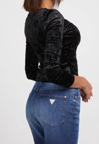Schwarzes Samt-Oberteil mit langen Ärmeln und figurbetonter Passform, kombiniert mit blauen Jeans aus Denim, die über ein dreieckiges Taschen-Detail und eine leichte Waschung verfügen.
