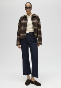 Vrouw met donkere zonnebril, bruin geruit jasje, crème hoodie, donkerblauwe wijd pijp jeans en bruine mocassins, staand tegen een effen achtergrond.