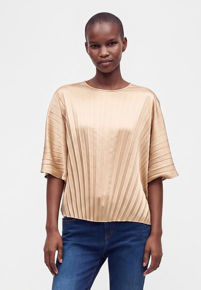 Femme portant un chemisier beige plissé à manches courtes et un jean bleu, debout sur un fond clair uni.