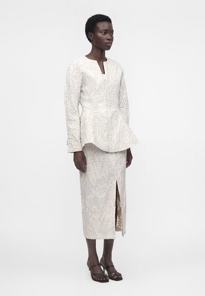 Γυναίκα στέκεται φορώντας λευκό υφασμάτινο μακρυμάνικο τοπ και ταιριαστή midi φούστα με σχίσιμο μπροστά, συνδυασμένα με μαύρα σανδάλια με τακούνι.