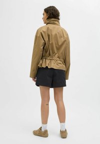 Chaqueta marrón ligera con cuello alto y cintura fruncida, combinada con pantalones cortos negros y zapatillas beige, contra un fondo neutro.