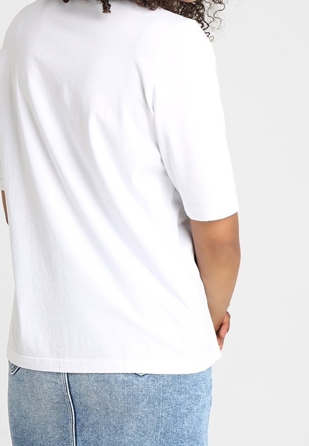 Osoba s kudrnatými vlasy, kterou má na sobě jednoduchou bílou košili s krátkým rukávem a světle modré džínsy, stojící s rukama za zády.