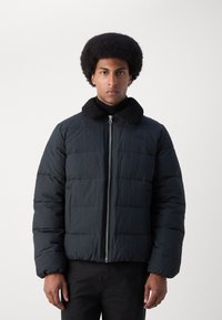 Sort pufferjakke med quiltet design, der har en tekstureret, sort shearlingkrave og frontlynlås.