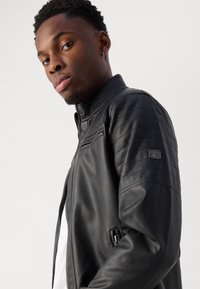 Jachetă de motocicletă din piele neagră cu prințe quilted la umeri, fermoar frontal, guler în picioare și un logo accent pe mânecă.