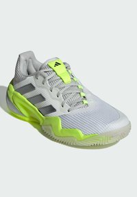 Zapatilla deportiva blanca con parte superior de malla gris, detalles en verde neón y tres franjas plateadas. Cuenta con una suela texturizada y área de puntera reforzada.