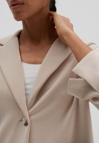 Světlý béžový blazer z hladké látky, s šálovým límcem, jednoduchým zapínáním na knoflík a ohrnutými rukávy. Jemný lesk a čisté linie.