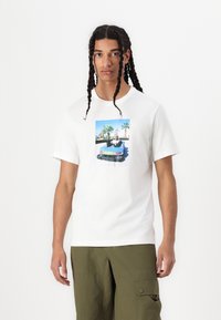 Tricou alb din bumbac cu un imprimeu grafic colorat care prezintă o persoană într-un go-kart. Designul include accente verzi, albastre și roșii.