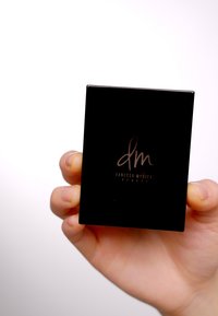 Svart rektangulær kompakt med en blank finish, med en subtil logo i mørkegrå. Holdt i en hånd, som fremhever størrelse og form.