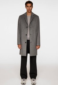 Gray wool overcoat with a straight cut, notched lapels, and five black buttons; worn over a light gray sweater and black trousers.