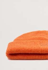 Oranssi neulepipo, jossa on ribbitekstuuri, taitettavan hihan ja kankaaseen brodeerattu hienovarainen logo.