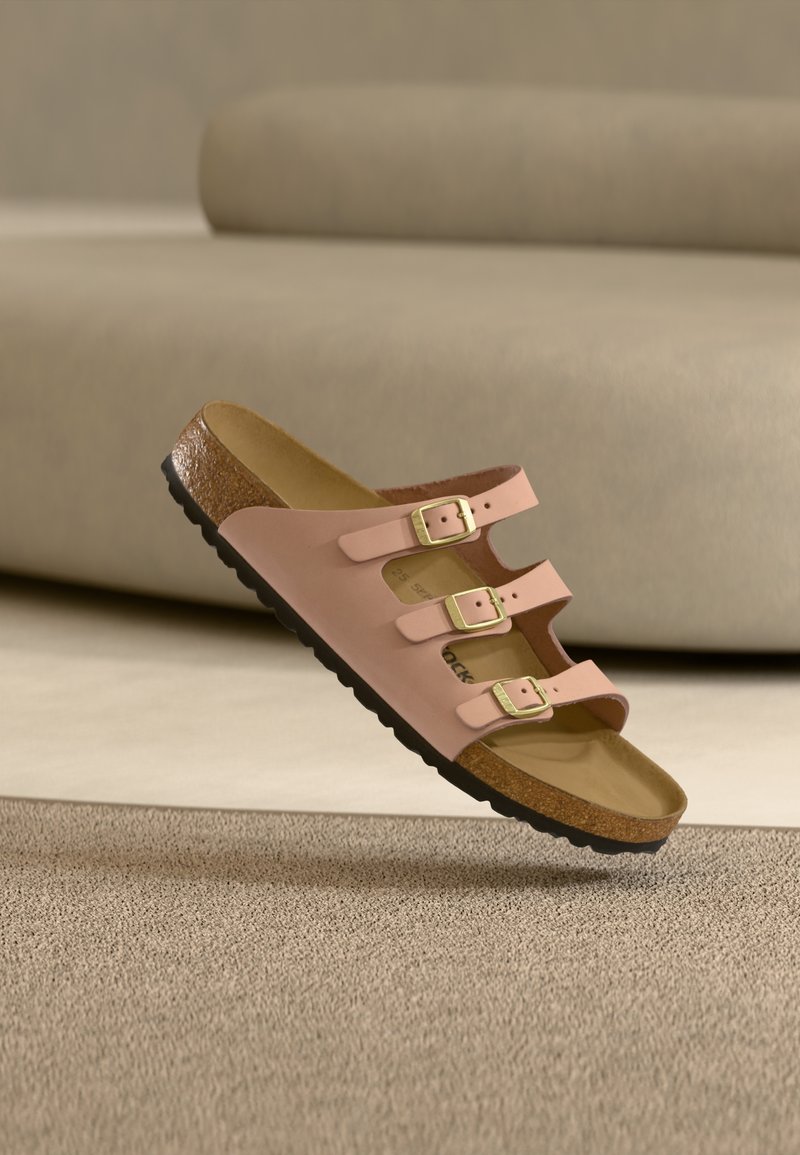 Ροζ ανοιχτό σανδάλι με τρεις χρυσές αγκράφες, φελλό πάτο και μαύρη σόλα που ακουμπά σε μπεζ χαλί κοντά σε μπεζ καναπέ.