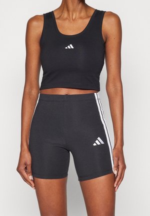 Kobieta w czarnym krótkim topie Adidas i dopasowanych czarnych szortach z białymi paskami i logo, stojąca na jednolitym tle.