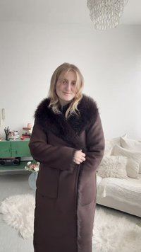 Noor naine siseruumides, kandes pikka pruunist mantlit tumeda karva kraega, seisab rohelise kapis ja valge diivani lähedal, millel on padjad.