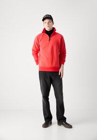 Crvena zip-up trenirka s dugim rukavima, uparena s crnim trapericama i crnim tenisicama. Na glavi se nosi crna kapa s logom "BOSS".