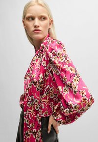 Blumenbluse mit rosafarbenem Hintergrund, langen Puffärmeln und hohem Kragen. Sie zeichnet sich durch schwarze und beiger Blumen mit einer glatten Textur aus.