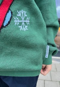 Grön stickad tröja med snöflinge-motiv i vitt, med ett rött och blått mönster. Inkluderar en etikett på fållen.