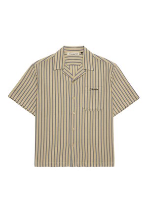 Femme portant une casquette marine, un t-shirt à manches courtes beige et gris rayé par-dessus un débardeur blanc brodé, et un pantalon gris clair avec une main dans la poche.