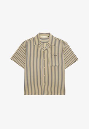 Frau trägt eine marineblaue Kappe, ein beige-grau gestreiftes Kurzarmshirt über einem weißen Tanktop mit Stickerei und hellgraue Hose, eine Hand in der Tasche.