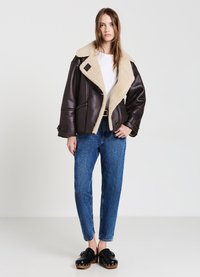 Giubbotto in pelle marrone con colletto in shearling crema, tasche con zip e chiusura a bottone. Indossato su una camicia bianca e abbinato a jeans blu.