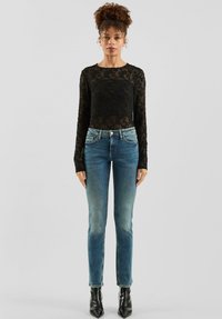 Czarna koronkowa bluzka z długim rękawem w połączeniu ze średnio niebieskimi dopasowanymi dżinsami. Jeansy mają delikatne wyblaknięcie oraz subtelne przeszycia, uzupełnione czarnymi butami na obcasie.