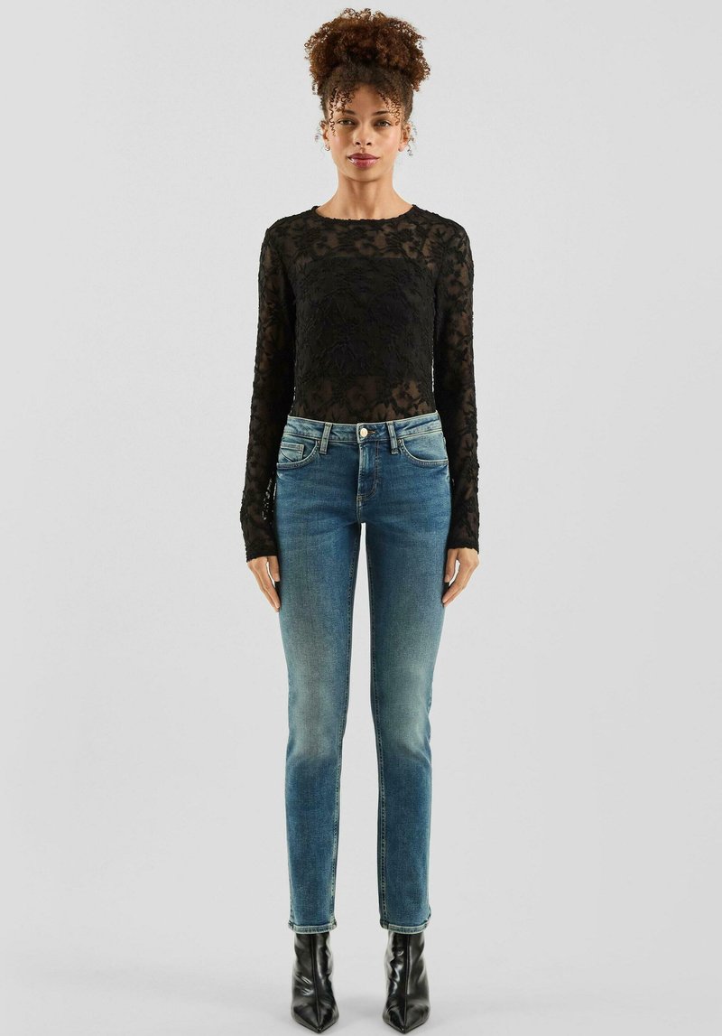 Czarna koronkowa bluzka z długim rękawem w połączeniu ze średnio niebieskimi dopasowanymi dżinsami. Jeansy mają delikatne wyblaknięcie oraz subtelne przeszycia, uzupełnione czarnymi butami na obcasie.