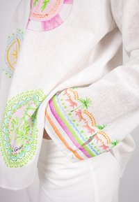 Weißes Leinenhemd mit bunten bestickten Mustern, darunter tropische Motive und Streifen in Pink, Grün und Orange.