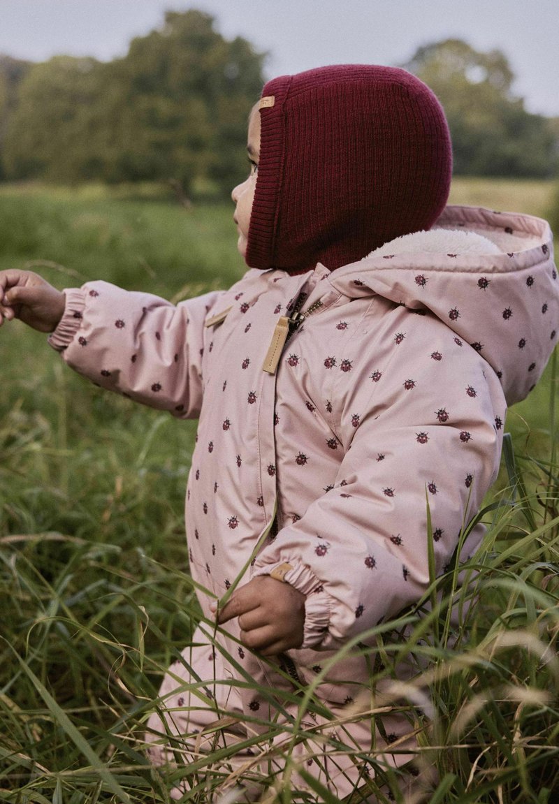 Roze pufferjack met bloemenpatronen, nep bontvoering en een geribde kastanjebruine muts. Een kind staat in hoog gras en strekt één hand uit.