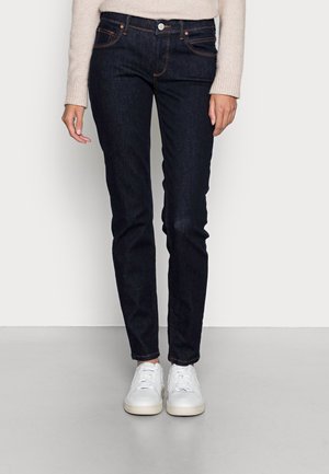 Woman wearing dark blue slim-fit jeans, beige sweater, and white sneakers standing on a light gray floor against a white wall.