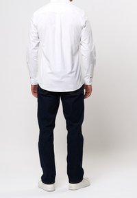 Biała koszula zapinana na guziki z długimi rękawami, pokazana od tyłu. W połączeniu z ciemnoniebieskimi dżinsami i białymi tenisówkami. Prosty krój i design.