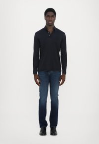 Ναυτικό πόλο μπλούζα με μακριά μανίκια και τρία κουμπιά, συνδυασμένο με σκούρες τζην παντελόνες και μαύρα παπούτσια. Το μοντέλο στέκεται μπροστά σε ουδέτερο φόντο.