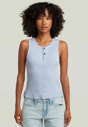 Donna con capelli ricci che indossa una canotta a coste azzurro chiaro con bottoni e jeans azzurri chiari, in piedi davanti a uno sfondo semplice.