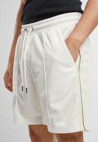 Pantaloni corti bianchi realizzati in tessuto liscio con vita elasticizzata, caratterizzati da un laccio e tasche laterali. Strisce verticali beige e nere sui lati.